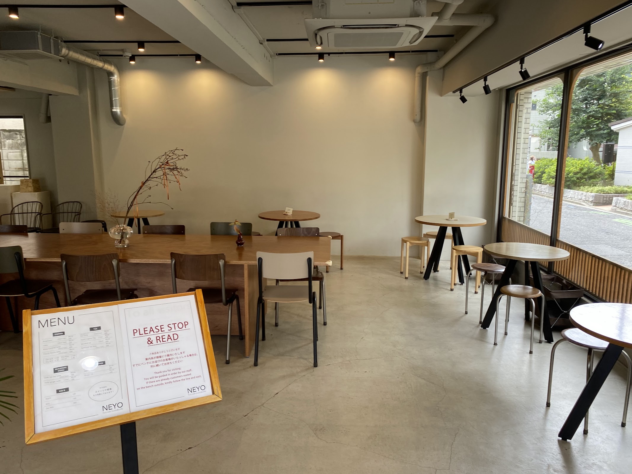 コンクリート床の広い店内に木製長テーブルと丸テーブルが並び、右手のアーチ窓から公園の緑が差し込むNEYOの全景