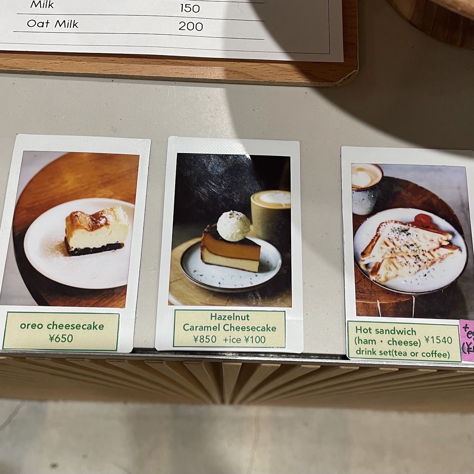 カウンター上に並ぶ3枚のインスタント写真風メニューカード。左からオレオチーズケーキ650円、ヘーゼルナッツキャラメルチーズケーキ850円、ホットサンド1540円の価格表示